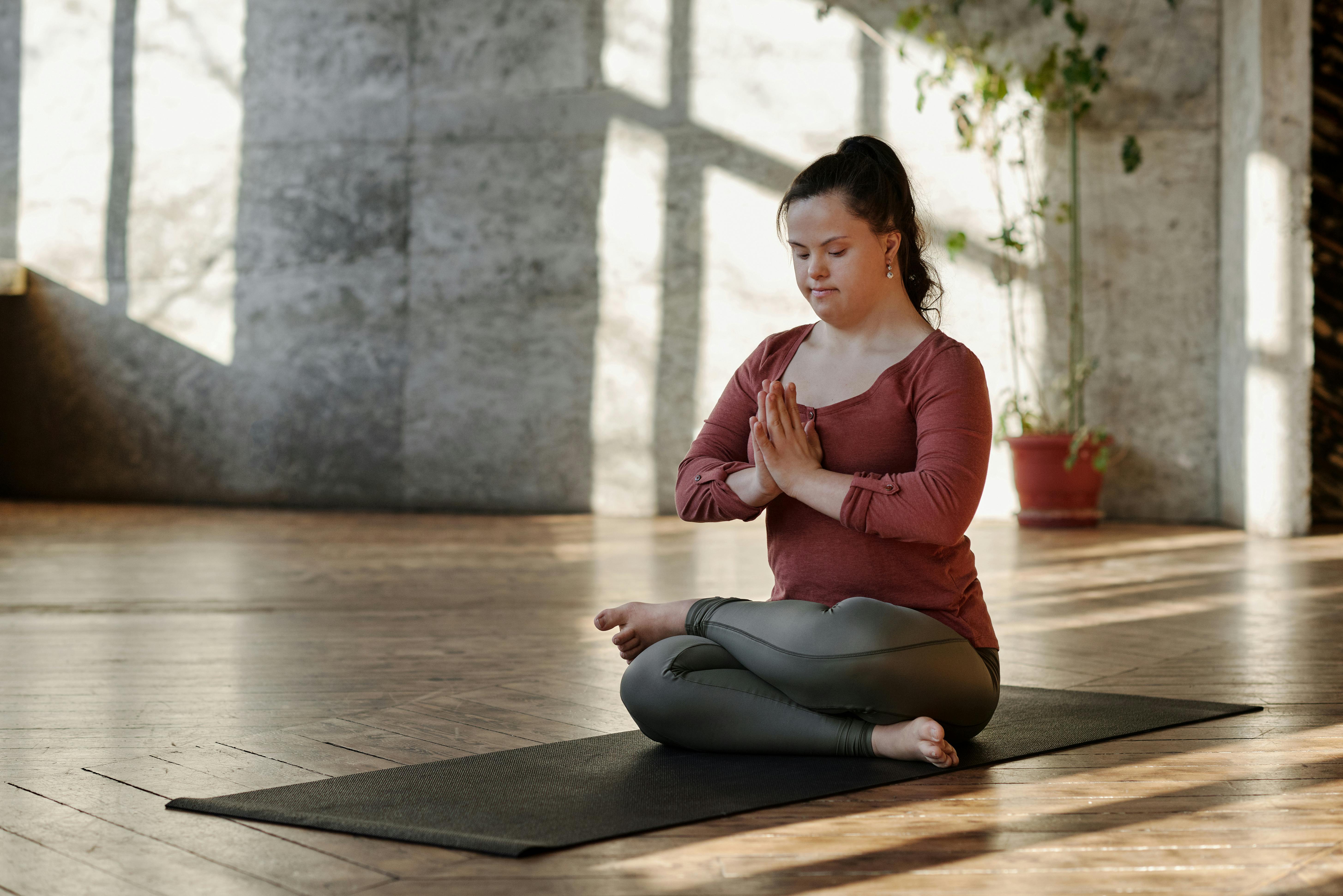 En kvinne med Downs syndrom sitter på en yogamatte på et tregulv innendørs. Hun sitter med bena i kryss og hendene samlet foran brystet. Hun har på seg langermet topp og treningstights, og håret er satt opp i hestehale. Bakgrunnen består av en vegg med steinflater og en potteplante. Lys og skygger faller på gulvet og veggen.