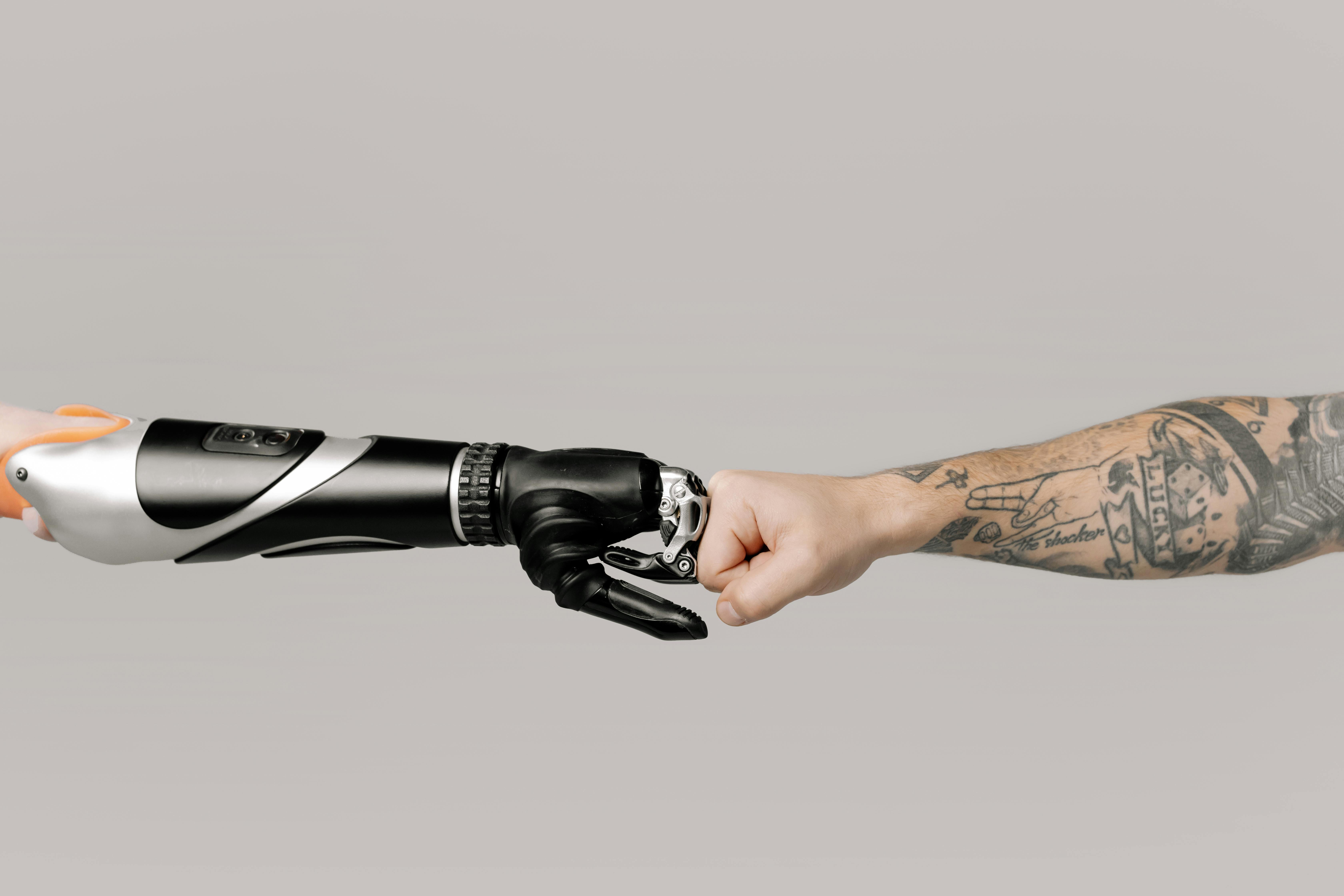 En protese laget av metall og plast kommer inn fra venstre og møter en tatovert underarm som kommer inn fra høyre i en fistbump mot en lys, nøytral bakgrunn.