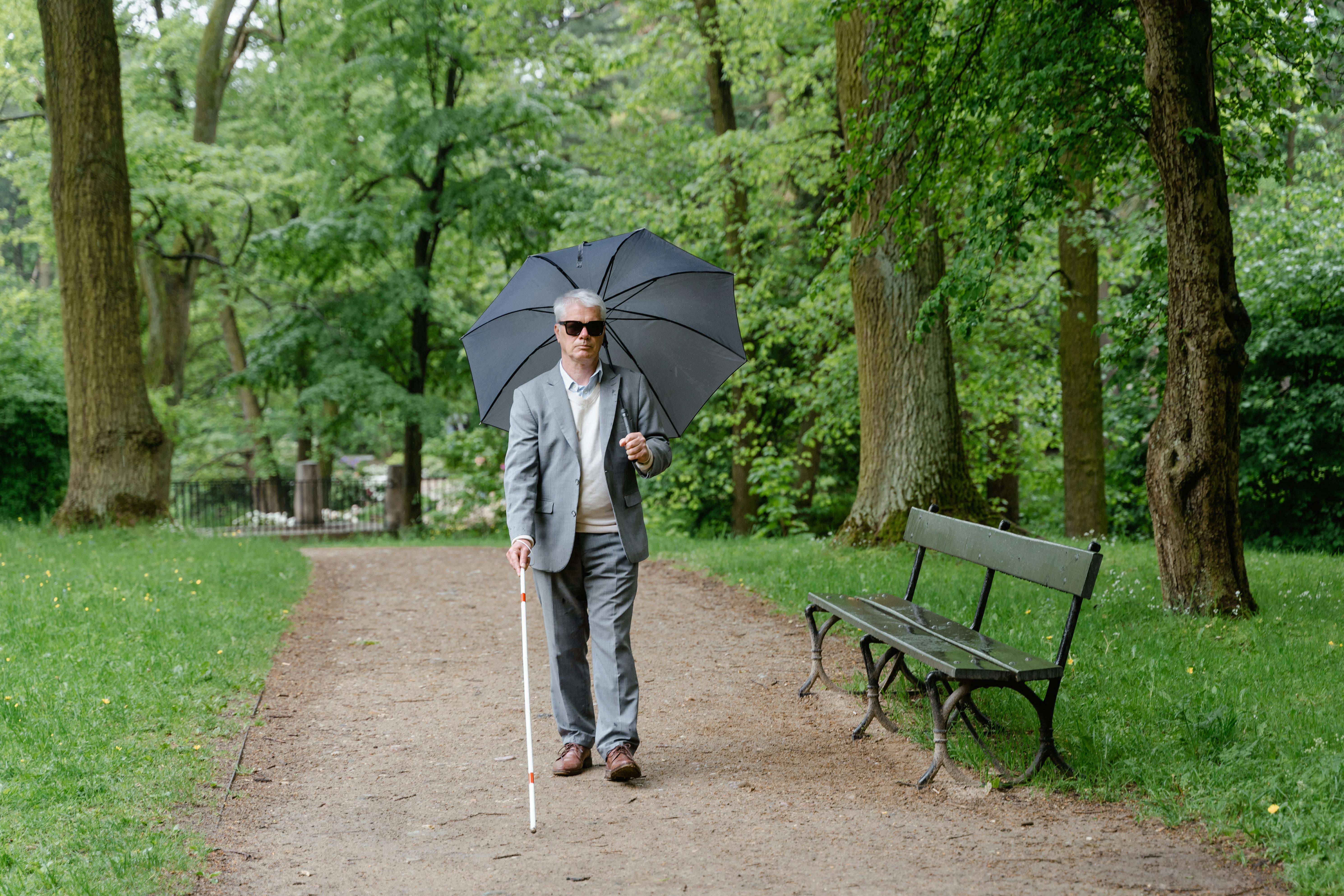 En eldre mann går på en grussti i en park. Han holder en hvit mobilitetsstokk i høyre hånd og en stor, mørkegrå paraply i venstre. Han har grått hår og er kledd i en lys skjorte, en beige genser og en grå dressjakke med matchende bukse, og han har på seg briller. Til høyre for stien står en mørkegrønn parkbenk, og parken rundt er fylt av høye trær og grønt gress med noen gule blomster spredt rundt.