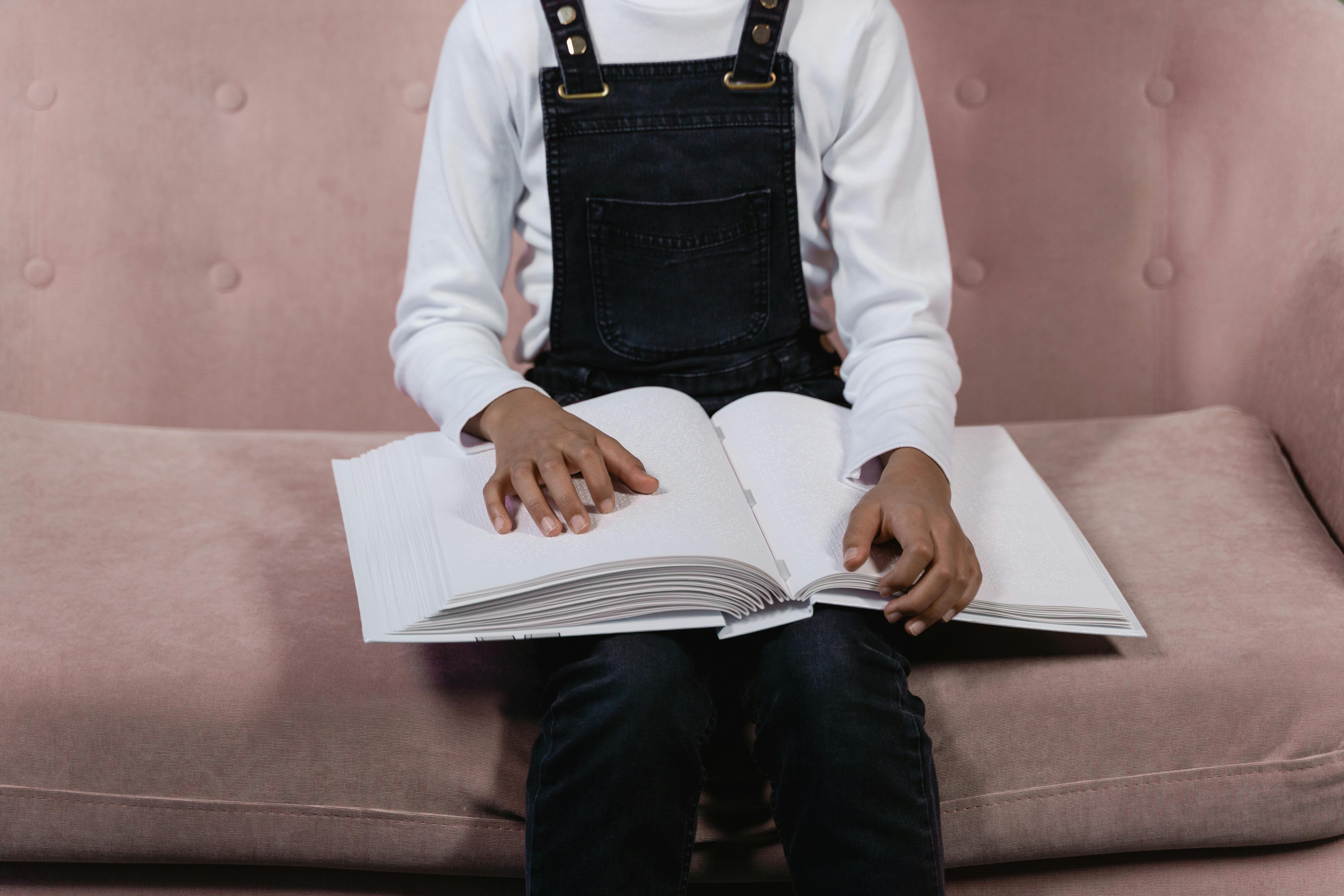 Et barn sitter på en rosa sofa og leser en stor bok med punktskrift. Barnet har på seg en hvit genser og mørk selebukse, og holder hendene på de åpne sidene mens fingrene følger punktskriften i boken.