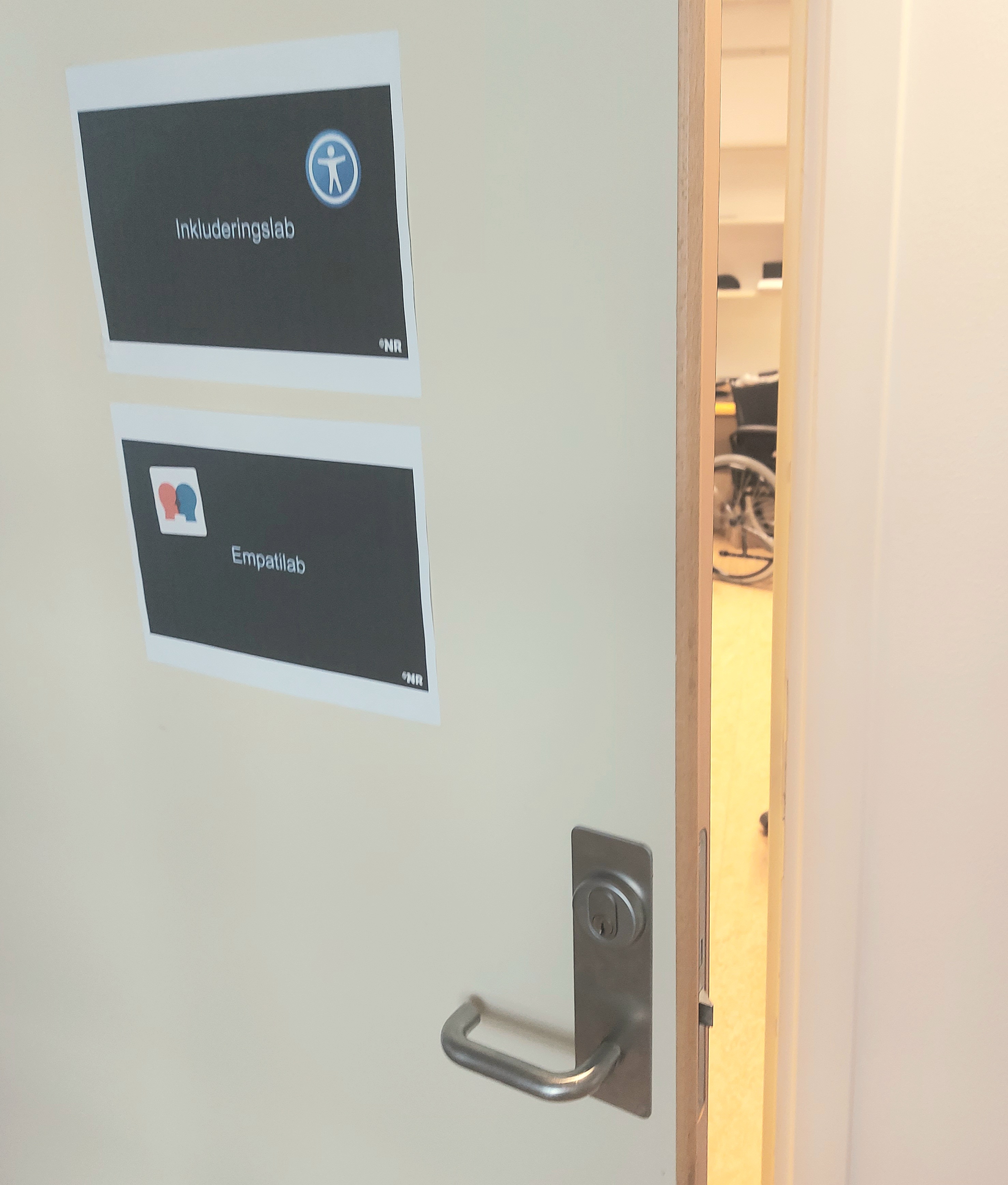 Dør med plakater det står "Inkluderingslab" og "Empatilab" på. Døren står på gløtt, og man kan skimte en rullestol inne.